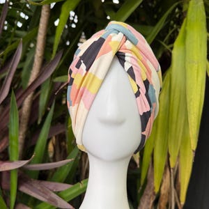 May include: A fabric turban headwrap featuring a knotted front detail and a multi-colored abstract geometric pattern in shades of dusty pink, mustard yellow, slate blue, black, and cream, displayed on a white mannequin head form.