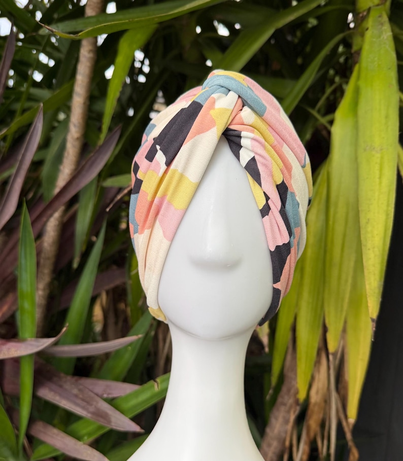 May include: A fabric turban headwrap featuring a knotted front detail and a multi-colored abstract geometric pattern in shades of dusty pink, mustard yellow, slate blue, black, and cream, displayed on a white mannequin head form.