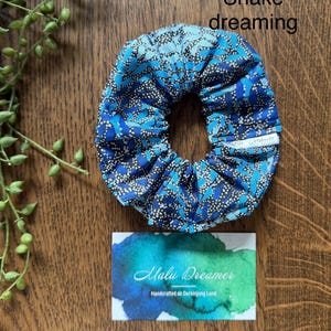May include: A blue and black patterned fabric scrunchie with a small white tag labeled Malu Dreamer. The text Snake dreaming is visible above the scrunchie, and a card with the text Malu Dreamer Handcrafted on Darkinjung Land is below.