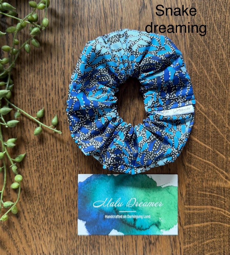 May include: A blue and black patterned fabric scrunchie with a small white tag labeled Malu Dreamer. The text Snake dreaming is visible above the scrunchie, and a card with the text Malu Dreamer Handcrafted on Darkinjung Land is below.