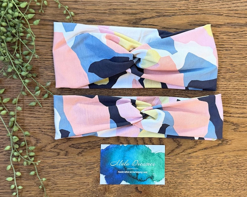 May include: Two fabric headbands featuring a twisted knot design and a multi-colored abstract pattern in shades of navy blue, light pink, pale yellow, and white. A rectangular card with a blue watercolor background and the text Malu Dreamer is included.