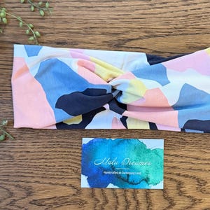 May include: A fabric headband featuring a twisted front design with an abstract geometric pattern in shades of pink, light blue, yellow, and dark navy blue. Below the headband is a rectangular card with a blue and green watercolor background.