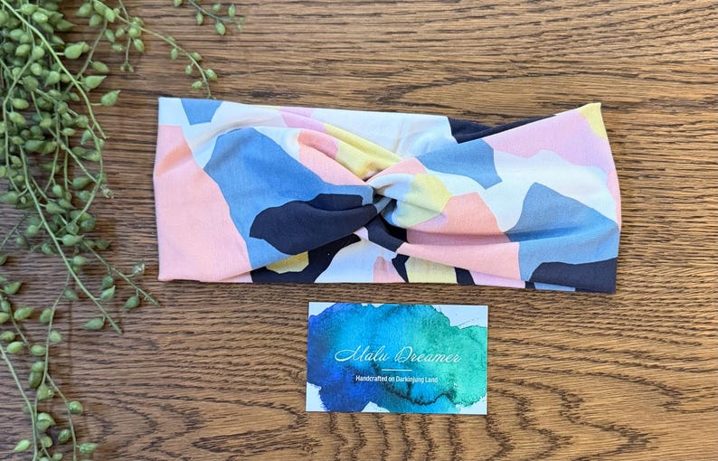 May include: A fabric headband featuring a twisted front design with an abstract geometric pattern in shades of pink, light blue, yellow, and dark navy blue. Below the headband is a rectangular card with a blue and green watercolor background.
