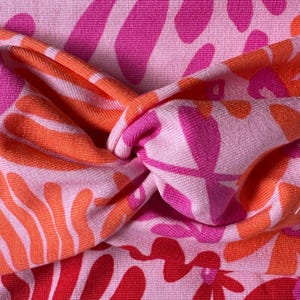 May include: A pink headband with a twisted knot design. The headband has a repeating pattern of orange, pink, and red floral shapes on a light pink background. The fabric appears to be soft and textured.