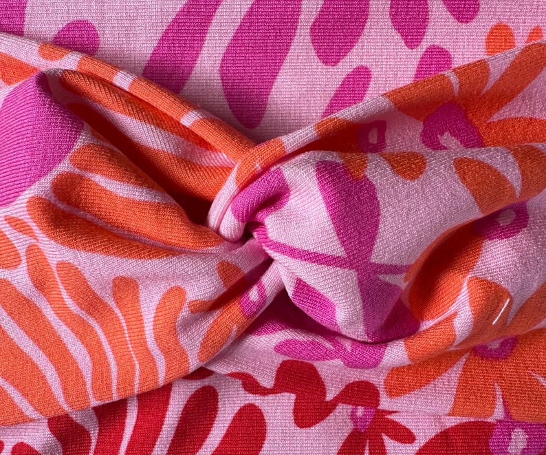 May include: A pink headband with a twisted knot design. The headband has a repeating pattern of orange, pink, and red floral shapes on a light pink background. The fabric appears to be soft and textured.