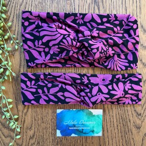 May include: Two fabric headbands featuring a black background with a vibrant purple floral and leaf pattern. Each headband has a twisted knot detail at the centre. A rectangular card with a blue and green watercolour design and the text Malu Dreamer.