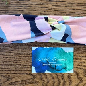 May include: A fabric headband featuring a twisted front and an abstract pattern of pink, navy blue, light blue, and pale yellow shapes. Below the headband is a rectangular card with a blue and green watercolor background and the text Malu Dreamer.