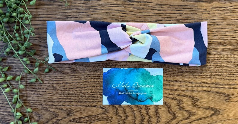 May include: A fabric headband featuring a twisted front and an abstract pattern of pink, navy blue, light blue, and pale yellow shapes. Below the headband is a rectangular card with a blue and green watercolor background and the text Malu Dreamer.