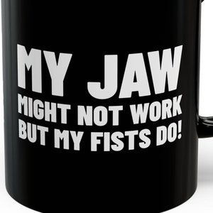 Puede incluir: Taza de café negra con texto blanco que dice "Mi mandíbula puede que no funcione, ¡pero mis puños sí!"