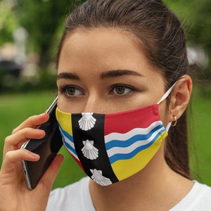 May include: A woman wearing a face mask with the flag of the Isle of Wight. The mask is black, yellow, red, and blue with white seashells. She is talking on a cell phone.
