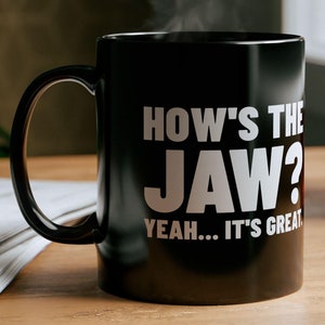 Puede incluir: Taza de café negra con texto blanco que dice "How's the Jaw? Yeah... It's Great."