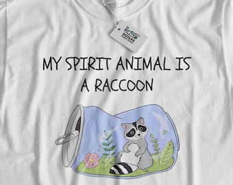 Op de afbeelding: Wit T-shirt met de tekst "Mijn spirit dier is een wasbeer" en een cartoon wasbeer in een blauwe blik.