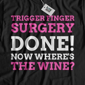 May include: Black t-shirt with the text "Trigger Finger Surgery Done! Now Where's The Wine?" in white and pink lettering.