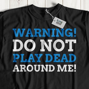 May include: Black t-shirt with white text that reads "WARNING! DO NOT PLAY DEAD AROUND ME!" in blue letters.