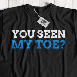 May include: Black t-shirt with the text "YOU SEEN MY TOE?" in white and blue lettering.