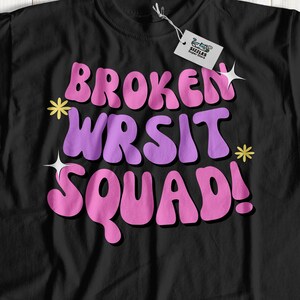 May include: Black t-shirt with a pink and purple graphic that says "BROKEN WRIST SQUAD!" with stars around the text.