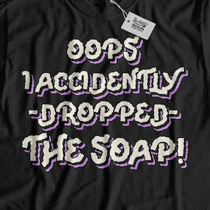 May include: Black t-shirt with a white graphic that says "Oops I accidentally dropped the soap!" in a cartoon font with purple outlines.