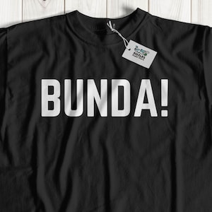 Op de afbeelding: Zwart t-shirt met het woord "BUNDA!" gedrukt in grote, witte hoofdletters. Het shirt heeft een ronde hals en korte mouwen. Een label is aan de kraag bevestigd. Het shirt wordt gepresenteerd op een licht houten oppervlak.