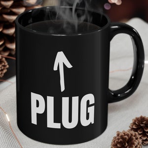 Puede incluir: Taza de café de cerámica negra con una flecha blanca apuntando hacia arriba y la palabra "PLUG" en letras blancas.
