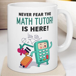 May include: White ceramic mug with a green calculator character carrying a suitcase. The mug has the text "Never fear the math tutor! Is here!" printed on it.