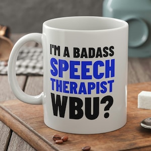 May include: White ceramic mug with a black and blue design. The mug reads "I'm a badass speech therapist WBU?"