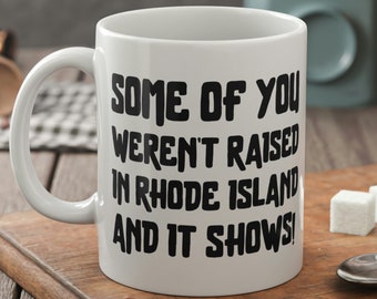 May include: White ceramic coffee mug with black text that reads "Some of you weren't raised in Rhode Island and it shows!"