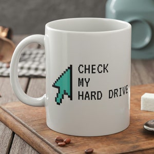 Pode incluir: Caneca de cerâmica branca com uma seta pixelada azul turquesa apontando para a direita e o texto "CHECK MY HARD DRIVE".