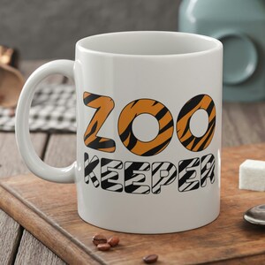 May include: White ceramic mug with a black and orange tiger stripe design. The text "ZOO KEEPER" is printed on the mug in black letters.