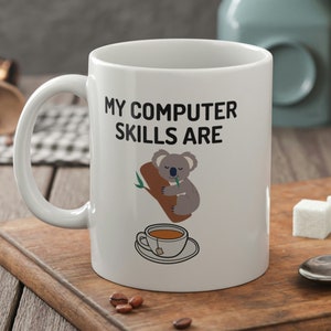 May include: White ceramic mug with a black graphic of a koala sleeping in a tree with a cup of coffee underneath. The text "My computer skills are" is above the koala.