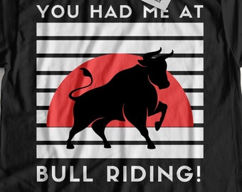 Puede incluir: Camiseta negra con un diseño gráfico blanco que presenta la silueta de un toro contra una puesta de sol roja con el texto "You had me at bull riding!"