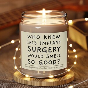 May include: A brown soy candle with a white label that reads "WHO KNEW IRIS IMPLANT SURGERY WOULD SMELL SO GOOD?"  Sizzle SuperStore // 9oz Eco-Friendly Soy Candle