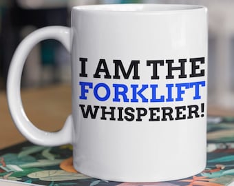 May include: White ceramic mug with a black and blue design that reads "I AM THE FORKLIFT WHISPERER!" The mug is sitting on a stack of books with a floral design.