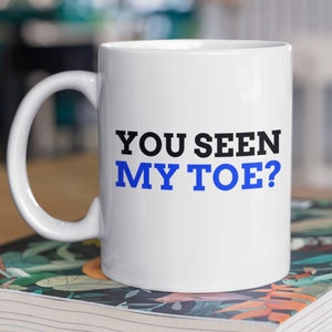 May include: White ceramic mug with a black and blue design. The text on the mug reads "YOU SEEN MY TOE?"