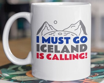 May include: White ceramic mug with a black and grey mountain illustration and the text "I must go Iceland is calling!" in blue and red. The mug is sitting on a stack of books with a floral pattern.