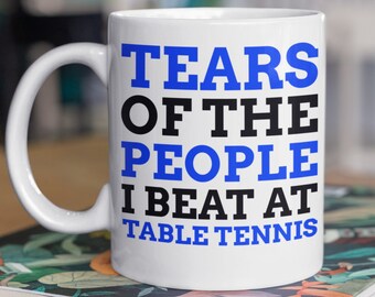 May include: White ceramic coffee mug with a blue and black design that reads "Tears of the People I Beat at Table Tennis".