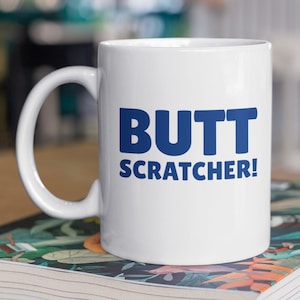 Puede incluir: Taza de cerámica blanca con una inscripción azul que dice "BUTT SCRATCHER!"