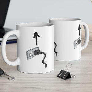 Puede incluir: Dos tazas de cerámica blanca con ilustraciones negras de un enchufe y una flecha apuntando hacia arriba. El texto "Plug it in" está escrito en la taza.