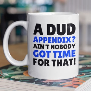 Peut inclure: Une tasse en céramique blanche avec un motif noir et bleu. La tasse porte l'inscription "A Dud Appendix? Ain't Nobody Got Time For That!"