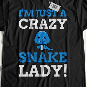 Op de afbeelding: Zwart T-shirt met de tekst "I'M JUST A CRAZY SNAKE LADY!" in wit en blauw. Een cartoon blauwe slang is gecentreerd onder het woord "CRAZY". Het shirt wordt gepresenteerd op een licht houten oppervlak.