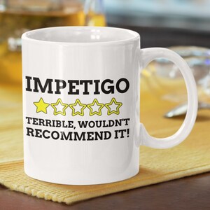May include: White ceramic coffee mug with a black and yellow design. The mug says "Impetigo" with a one-star rating and the text "Terrible, wouldn't recommend it!"