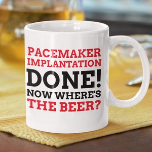 Puede incluir: Taza de café de cerámica blanca con texto negro que dice "Pacemaker Implantation Done! Now Where's The Beer?" El texto "Done!" está en rojo.