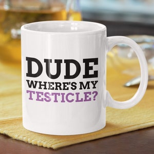 May include: White ceramic coffee mug with a black and purple design. The mug has the text "DUDE WHERE'S MY TESTICLE?" printed on it.