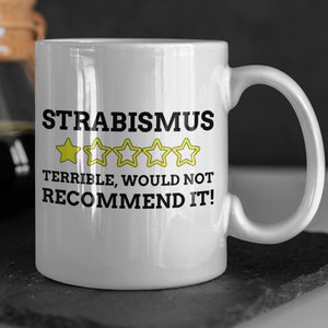 Puede incluir: Taza de café de cerámica blanca con un diseño en negro y amarillo. La taza tiene el texto "Strabismus" y una calificación de una estrella con el texto "Terrible, no lo recomendaría!"