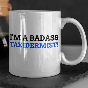Könnte beinhalten: Weiße Keramik-Kaffeetasse mit einem schwarzen und blauen Design. Die Tasse trägt die Aufschrift "I'm a badass taxidermist!"
