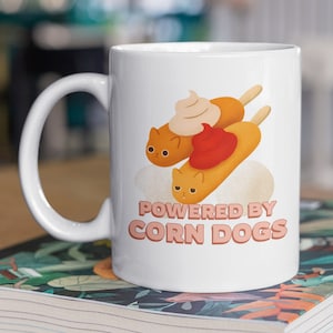 May include: White ceramic mug with a cartoon illustration of corn dogs with mustard and ketchup. The text "POWERED BY CORN DOGS" is printed in pink on the mug.