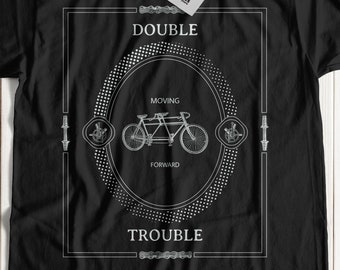May include: Black t-shirt with a white graphic design featuring a tandem bicycle with the text "DOUBLE TROUBLE" and "MOVING FORWARD" in a circle.