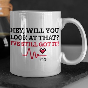 May include: White ceramic mug with a black and red graphic that reads "Hey, will you look at that? I've still got it!" with a red heart and a heartbeat line with the number 120 below it.