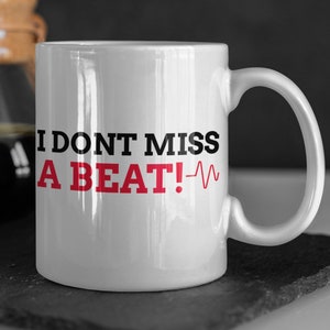 May include: White ceramic mug with a black and red graphic that says "I don't miss a beat!" with a red heartbeat symbol.