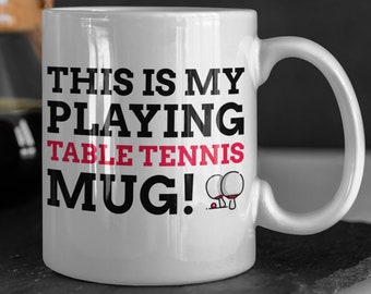 May include: White ceramic mug with black text that reads "This is my playing table tennis mug!" with a red and black illustration of two ping pong paddles and a ball.