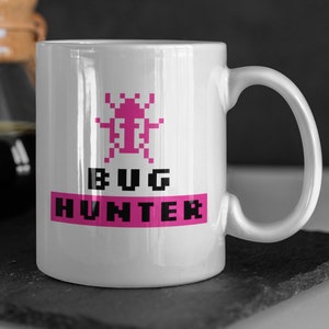 Puede incluir: Taza de cerámica blanca con un insecto pixelado rosa y el texto "BUG HUNTER" en negro.